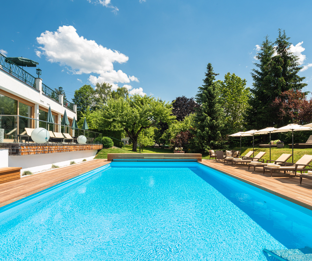Large outdoor swimming pool with sun loungers and parasols, surrounded by trees and a modern house.
