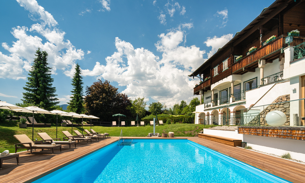 Luxushotel mit Swimmingpool, Liegestühlen und grünem Garten unter einem strahlend blauen Himmel mit Wolken.