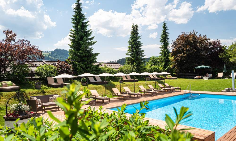 Liegestühle und Sonnenschirme an einem klaren Außenpool, umgeben von grünem Gras und hohen Bäumen an einem sonnigen Tag.