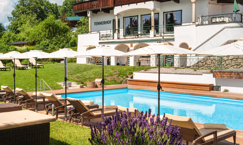 Außenpool mit Liegestühlen und Sonnenschirmen vor einem Hotelgebäude, umgeben von Grünflächen und Blumen.
