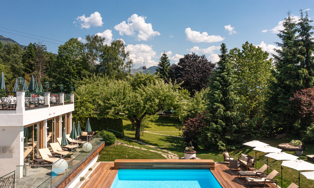 Sonniger Außenpool mit Liegestühlen, umgeben von Bäumen und einem Garten, neben einem modernen weißen Gebäude.