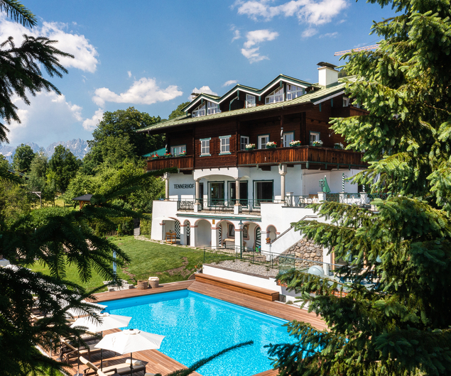 Großes Haus im Chalet-Stil mit Balkonen und Blick auf einen blauen Swimmingpool, umrahmt von üppigen grünen Bäumen.