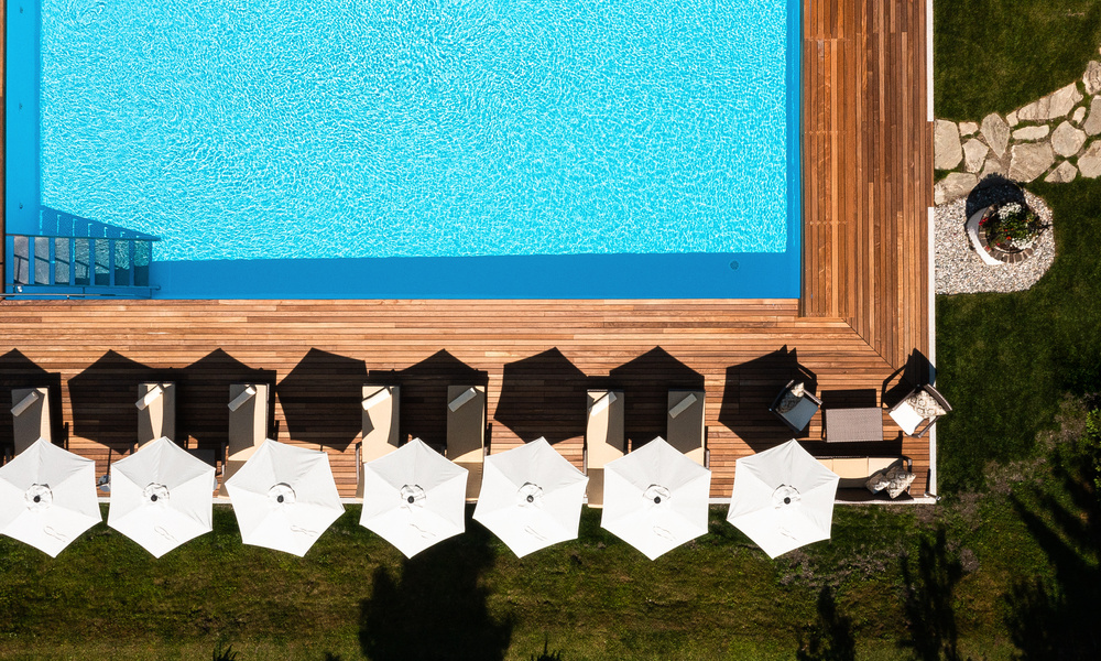Luftaufnahme eines Pools, einer Holzterrasse, weißer Sonnenschirme, Liegestühle und eines angelegten Gartens.