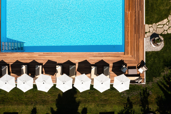 Luftaufnahme eines Pools, einer Holzterrasse, weißer Sonnenschirme, Liegestühle und eines angelegten Gartens.