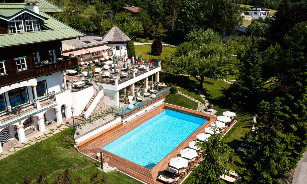 Luftaufnahme eines Hotels mit einer Dachterrasse und einem rechteckigen, von Grün umgebenen Außenschwimmbad.