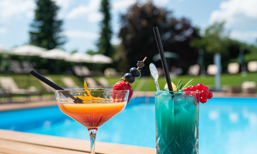 Zwei farbenfrohe Cocktails an einem Pool, mit Garnierungen, Bäumen und Liegestühlen im Hintergrund.