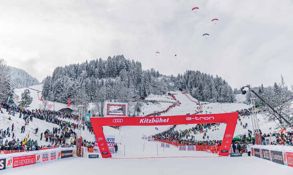 Eine verschneite Ziellinie eines Skirennens mit Menschenmassen, Bannern und Skifahrern, die einen bewaldeten Berghang hinunterfahren.
