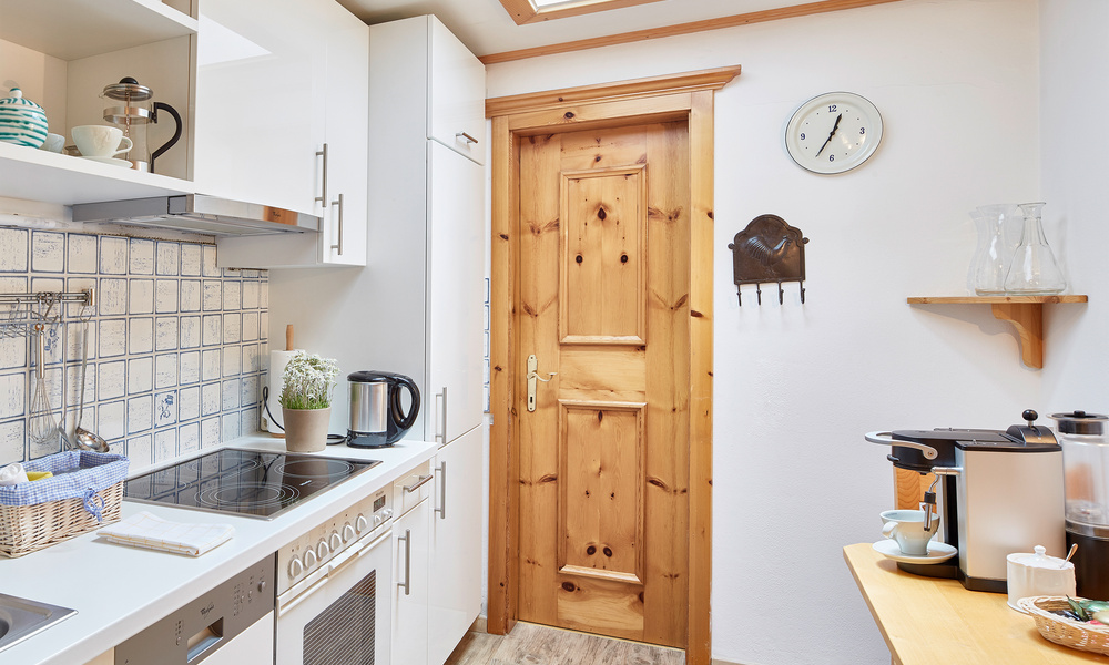 Gemütliche weiße Küche mit Holztür, Uhr, Geräten, Regalen und gefliestem Aufsatz.