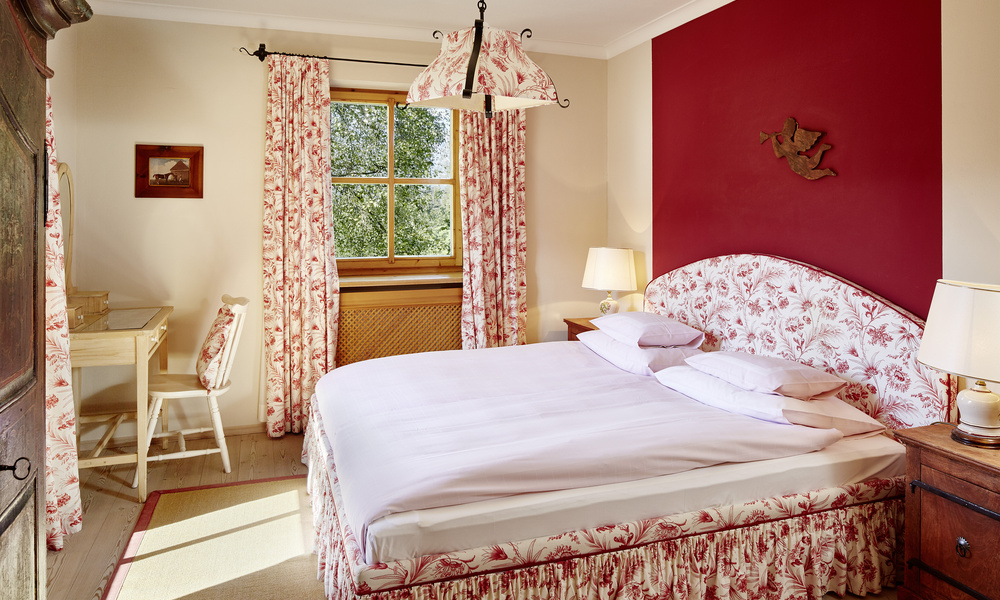 Gemütliches Schlafzimmer mit Doppelbett, floralem Muster, roter Akzentwand, Schreibtisch und Fenster mit Vorhängen.