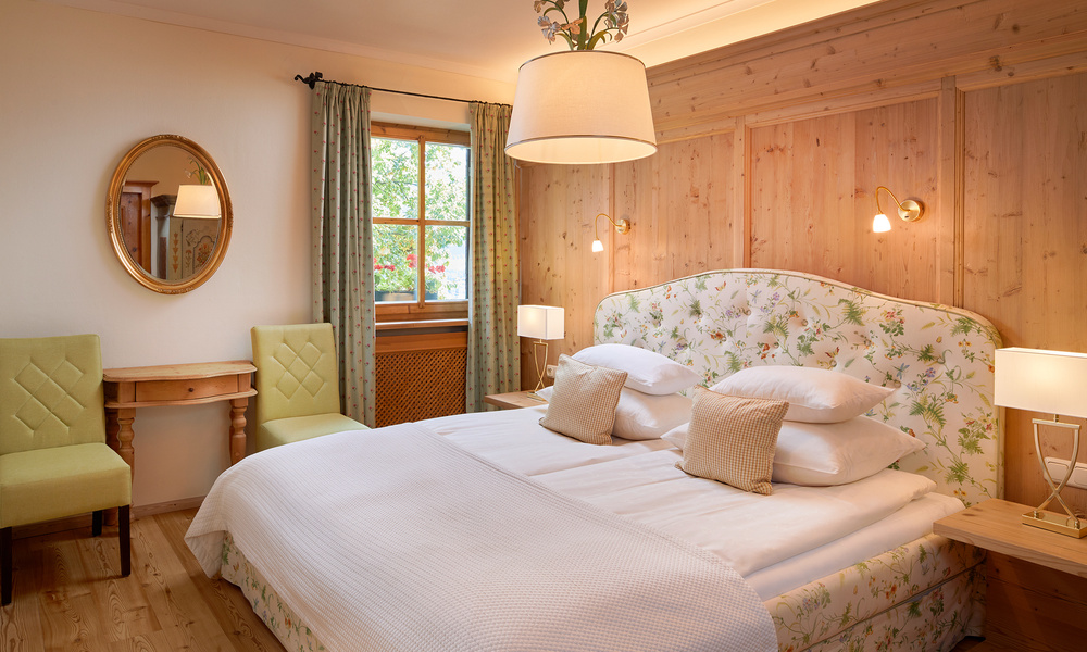 Gemütliches Schlafzimmer mit Blumendekor, Doppelbett, Holzwänden, grünen Stühlen und einem Fenster mit grünen Vorhängen.