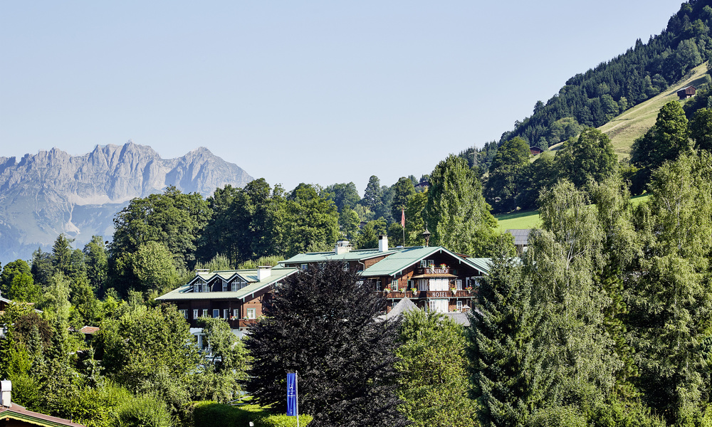 Ein Gebäude im Chalet-Stil, umgeben von Bäumen mit Bergen und grünen Hügeln im Hintergrund.