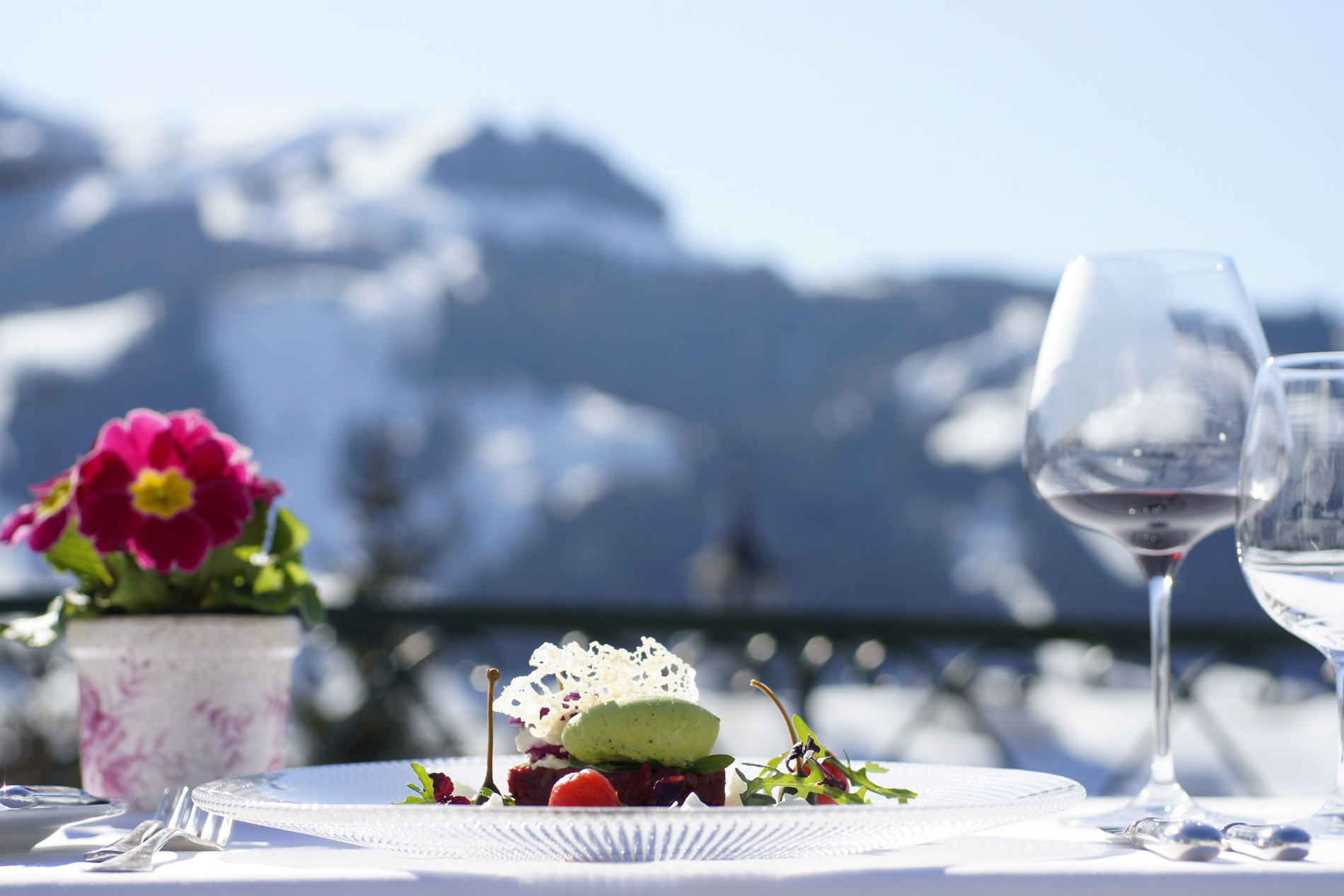 Gourmetteller und Weinglas auf einem Tisch mit Blumen, vor einer verschneiten Bergkulisse.