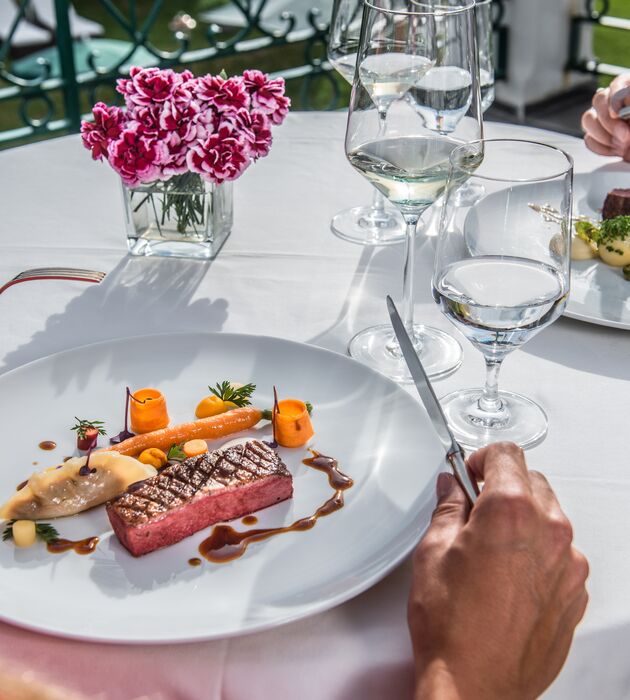 Eine Person schneidet ein Steak mit Gemüse auf einem weißen Teller an einem eleganten Esstisch im Freien an.