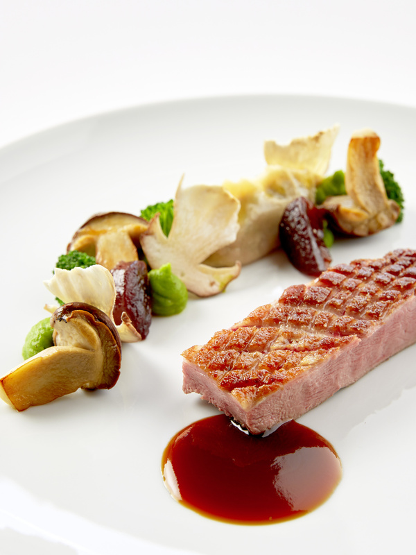 Ein Gourmetgericht mit gebratenem Fleisch, Soße, Pilzen, Brokkoli und Gemüsebeilagen auf einem weißen Teller.