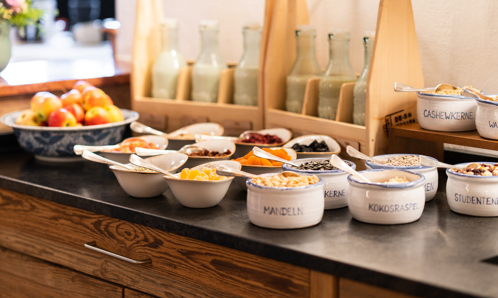 Schüsseln mit verschiedenen Müslis, Nüssen und Obstbelägen auf einer Theke neben Flaschen und einer Schale mit Äpfeln.