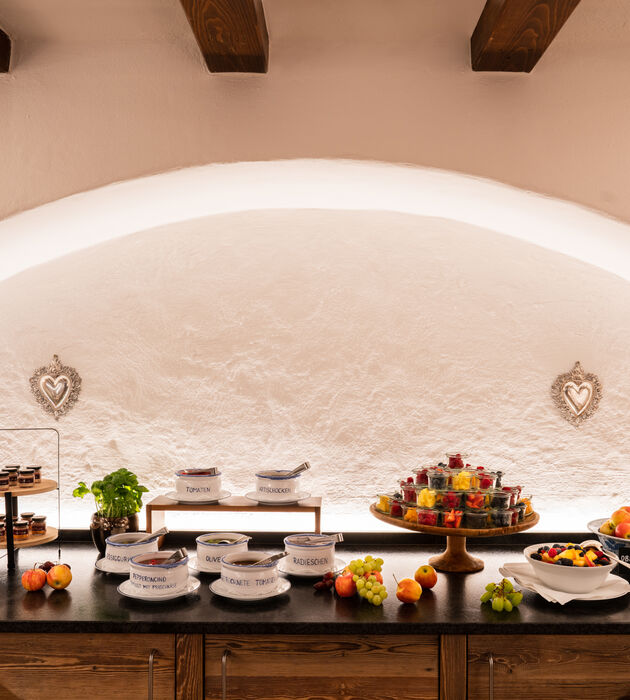 Buffet-Tisch mit Schalen mit Obst, Desserts und beschriftetem Geschirr vor einer weißen, gewölbten Wand.