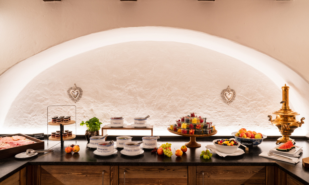 Buffet-Tisch mit Schalen mit Obst, Desserts und beschriftetem Geschirr vor einer weißen, gewölbten Wand.