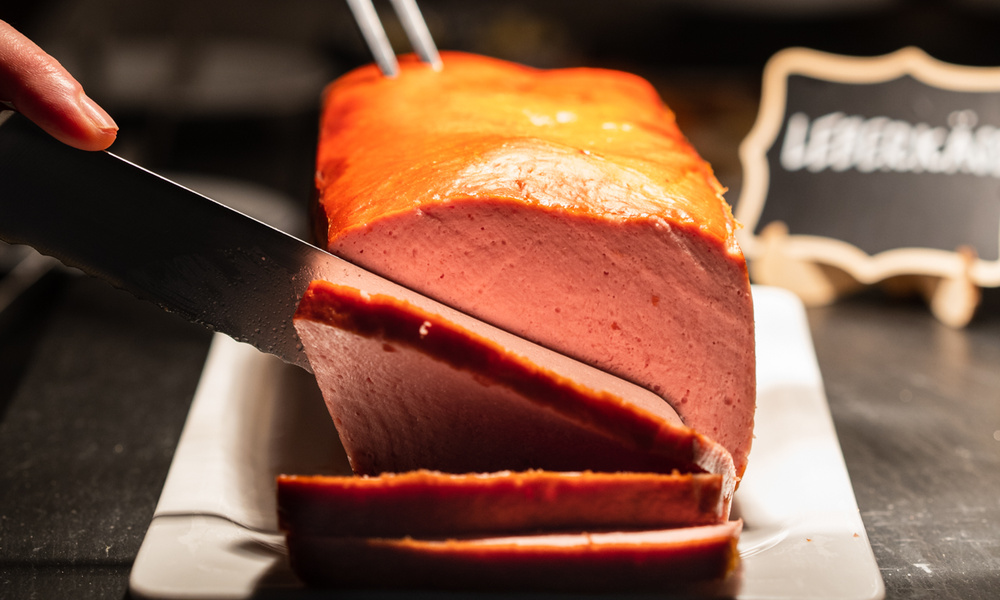 Eine Hand schneidet mit einem Messer und einer Zange einen Laib Leberkäse auf einem weißen Teller.