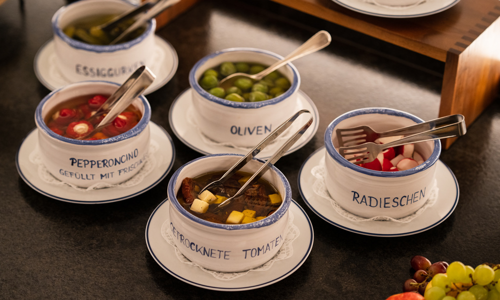 Fünf Schalen mit Servierbesteck, gefüllt mit verschiedenen eingelegten Gemüsesorten und in deutscher Sprache beschriftet, auf einer dunklen Arbeitsplatte.