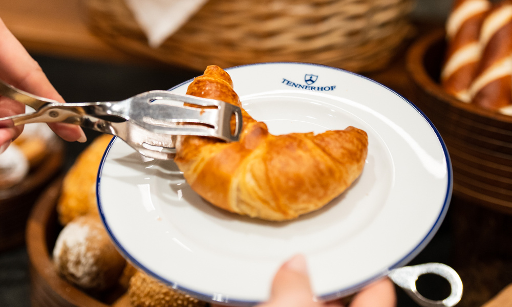 Ein Croissant wird mit einer Zange auf einen weißen Teller gelegt, im Hintergrund sind Brötchen zu sehen.