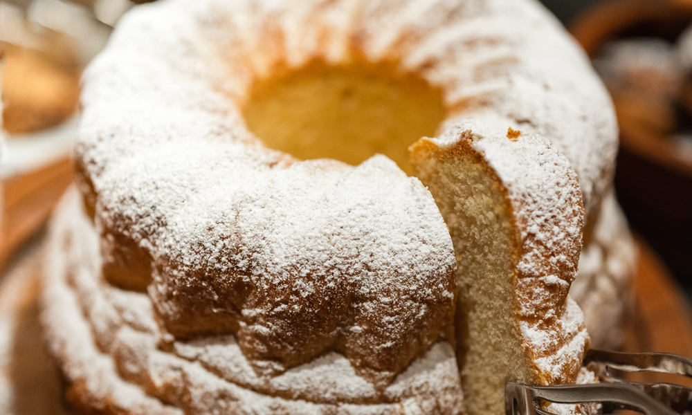 Ein runder, mit Puderzucker bestreuter Biskuit, aus dem eine Scheibe herausgeschnitten wurde, die seine weiche, fluffige Textur zeigt.