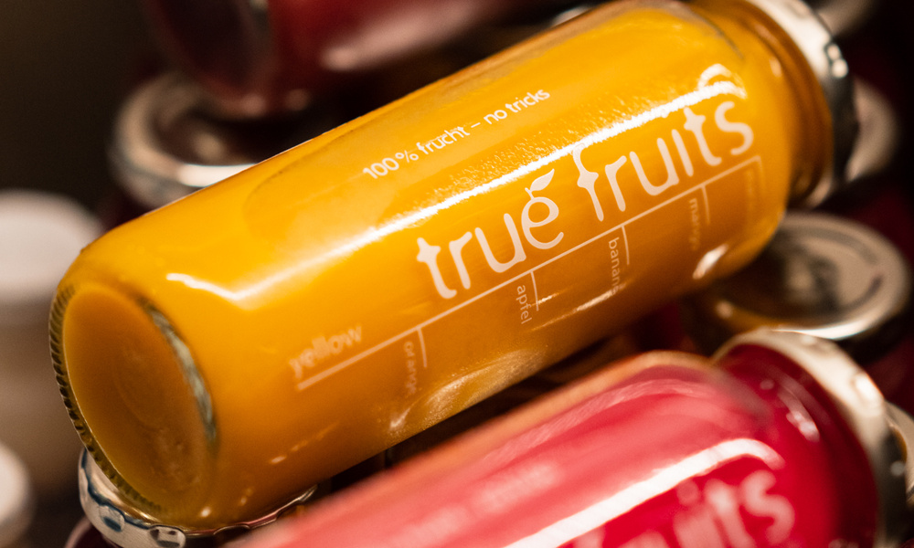 Eine Glasflasche mit einem gelben Smoothie aus echten Früchten liegt auf roten Smoothie-Flaschen.