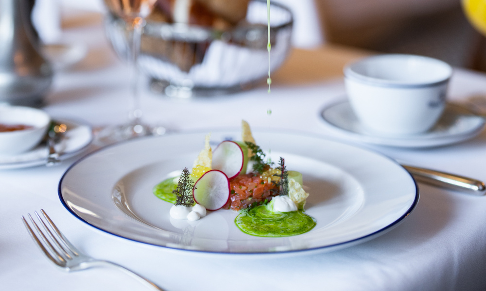 Eine Hand gießt grüne Soße auf einen Gourmet-Teller, garniert mit Radieschen und Kräutern auf einem weißen Teller.