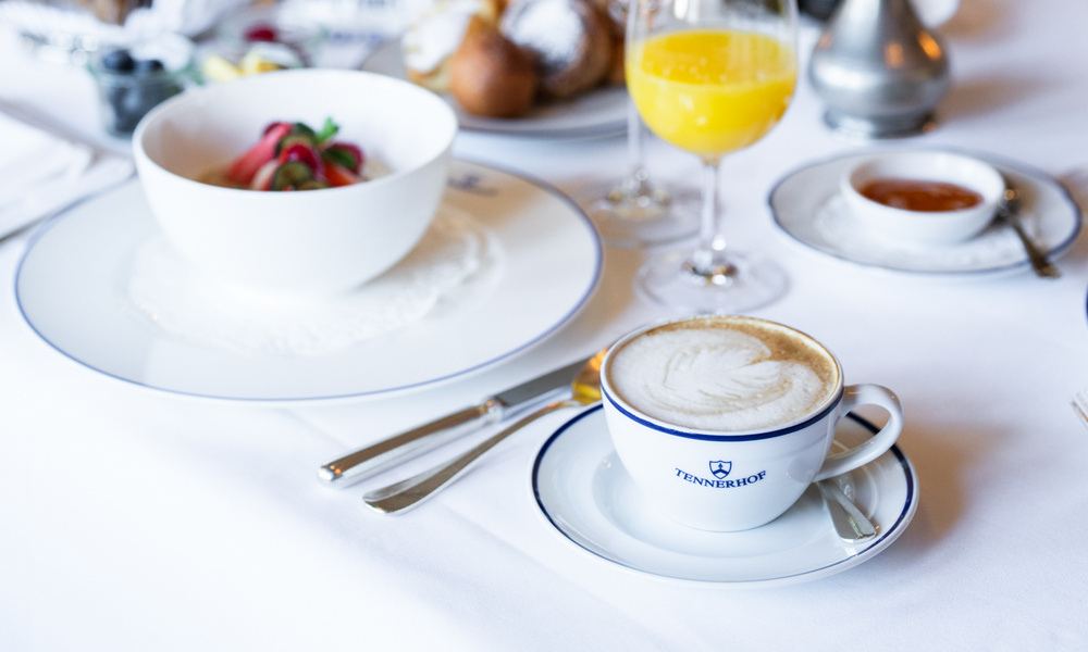 Ein Cappuccino, eine Obstschale und Orangensaft auf einem weißen Tischtuch, das für das Frühstück gedeckt ist.