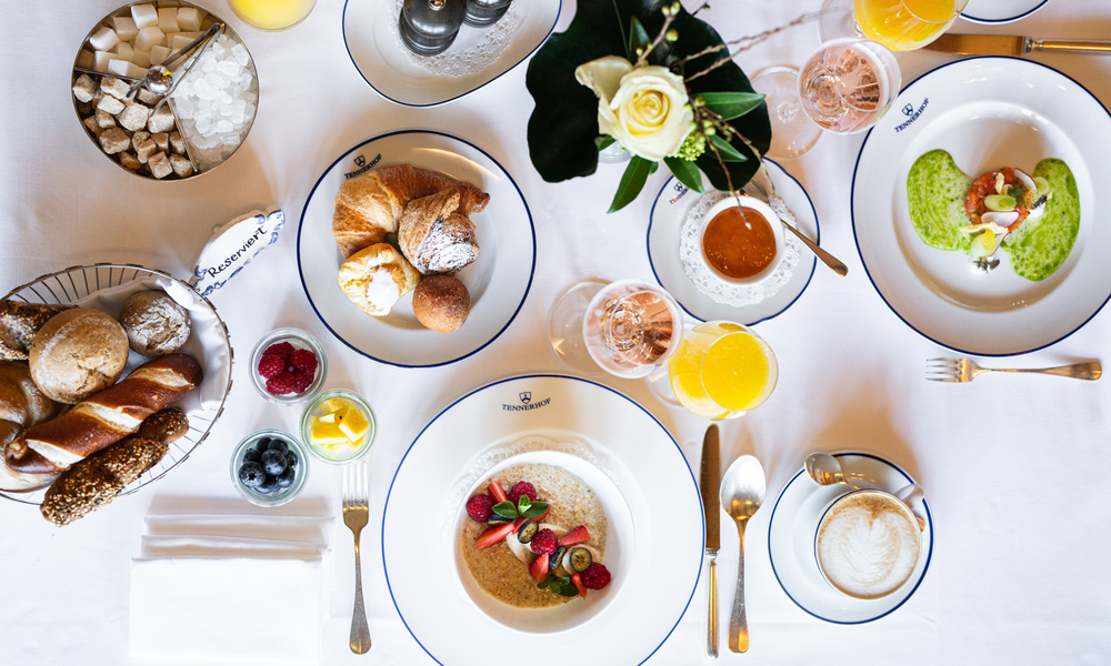 Eleganter Frühstückstisch mit Gebäck, Obst, Saft, Kaffee und dekorativen Blumen auf weißem Tischtuch.