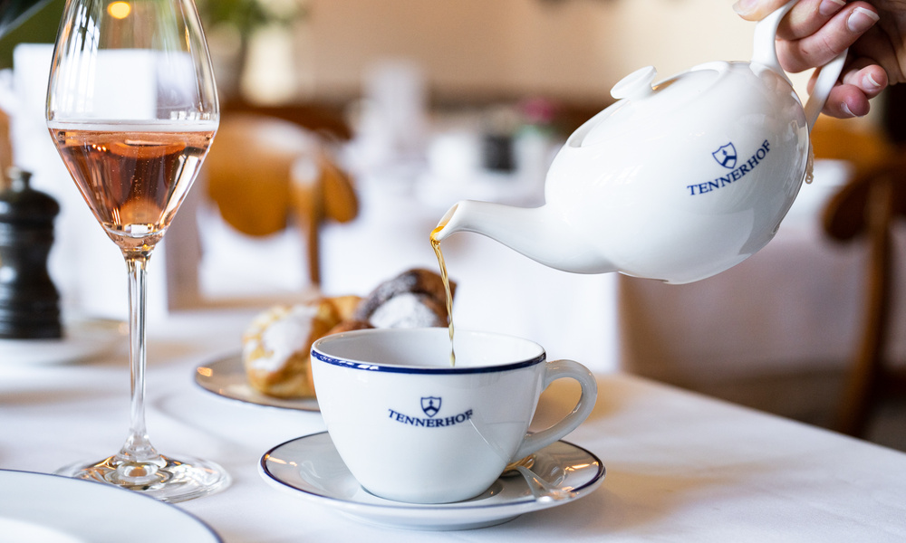 Eine Hand gießt Tee aus einer weißen Teekanne in eine passende Teetasse, daneben stehen ein Glas Roséwein und Gebäck.