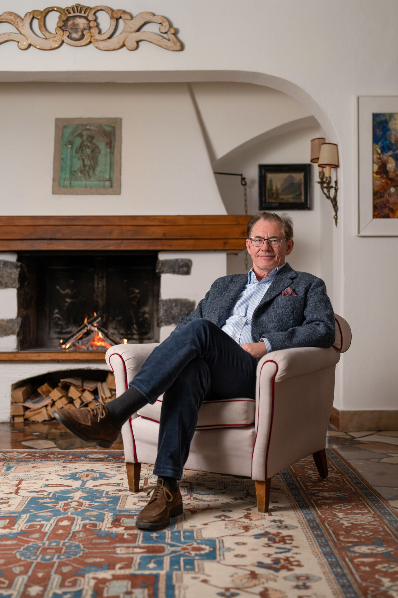 A smiling man in a blazer sits in an armchair in front of an illuminated fireplace in a comfortably furnished living room.