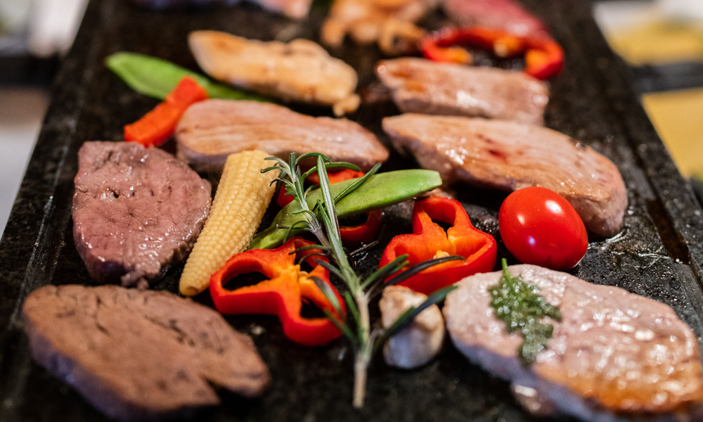 Verschiedene Fleisch- und Gemüsesorten, darunter Paprika und Kirschtomaten, die auf einem Grill oder heißen Stein gegart werden.