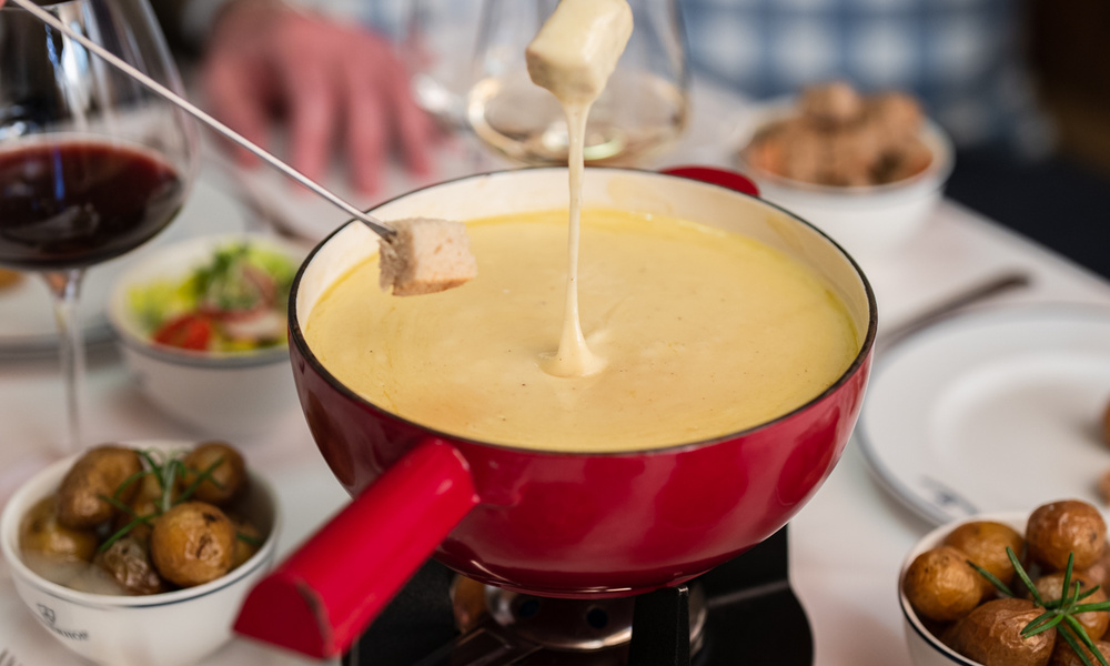 Zwei Personen tauchen Brot in einen Topf mit Käsefondue, umgeben von Schüsseln mit Kartoffeln und Gläsern mit Wein.