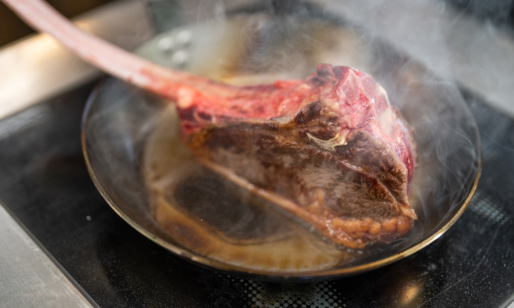 Ein großes Tomahawk-Steak brutzelt und dampft in einer heißen Pfanne auf dem Herd.