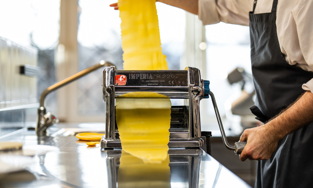 Person, die in einer Küche mit einer Nudelmaschine lange Blätter aus frischem Nudelteig ausrollt.