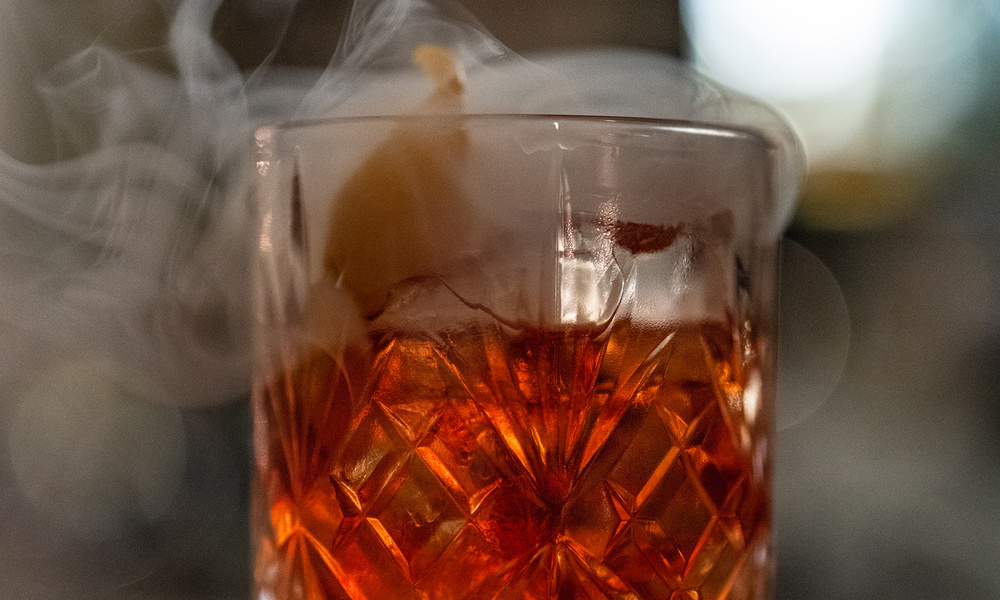 Ein rauchiger Cocktail in einem gemusterten Glas mit einer orangefarbenen Garnierung und wirbelndem Rauch obenauf.