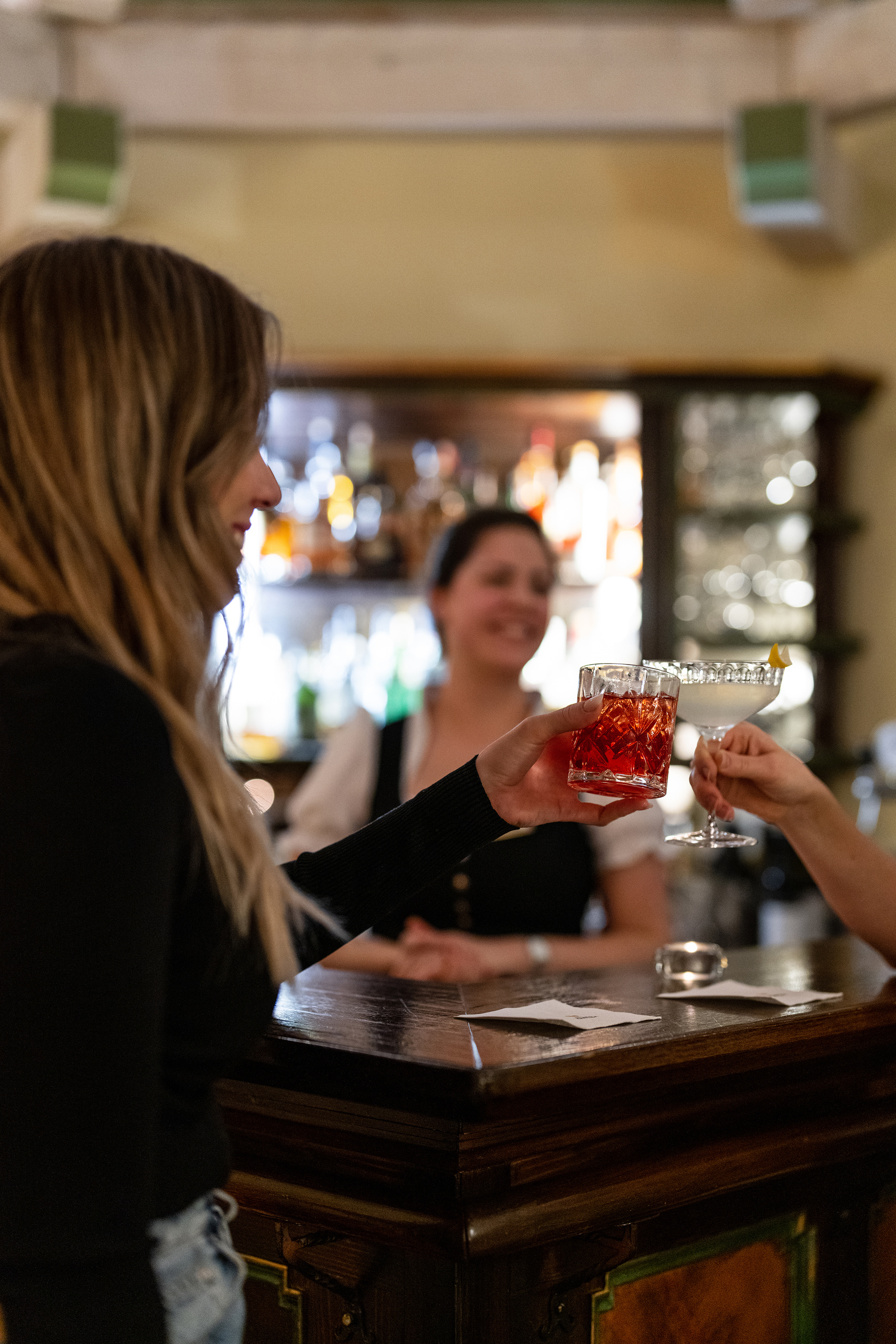 Two people toast their cocktail glasses at a bar with a smiling bartender in the background.