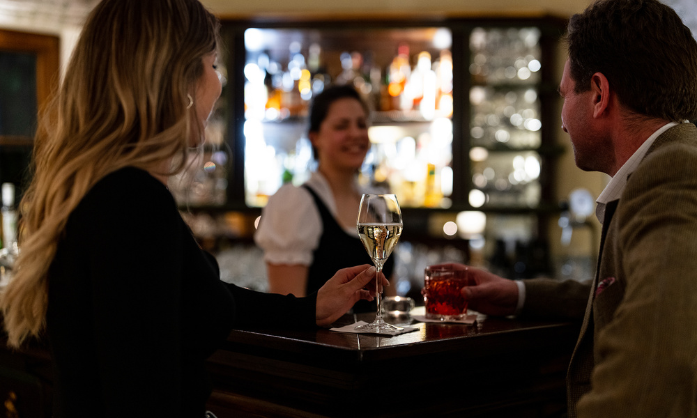 Zwei Personen mit Getränken unterhalten sich an einem Bartresen, während ein Barkeeper im Hintergrund lächelt.