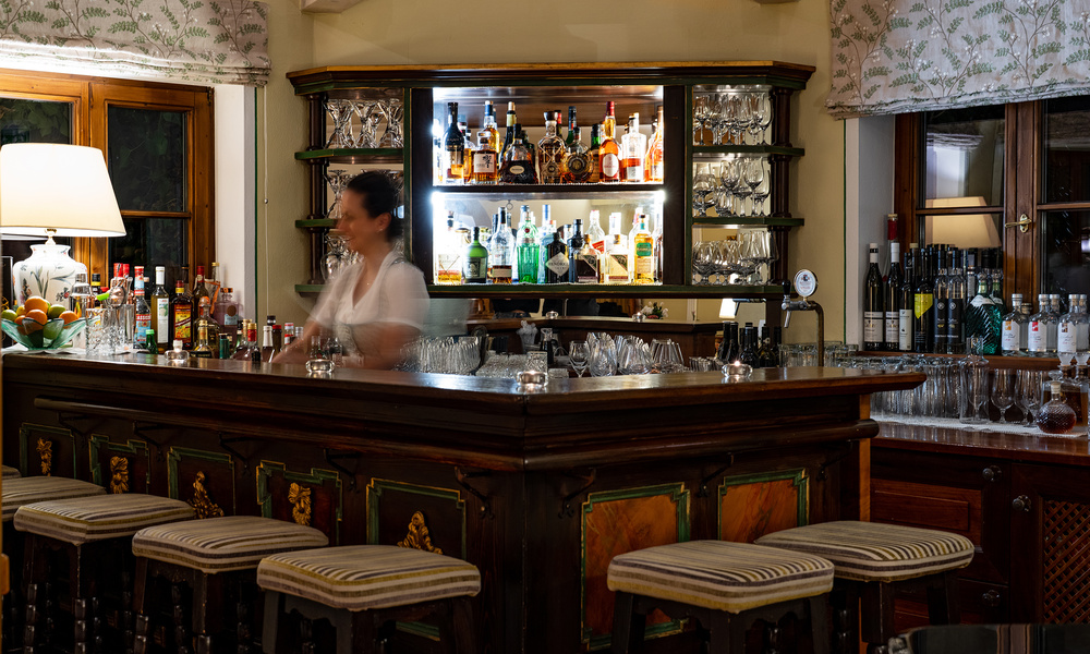 Ein Barkeeper steht hinter einer hölzernen Bar mit verschiedenen Flaschen und Gläsern in einem warm beleuchteten Raum.