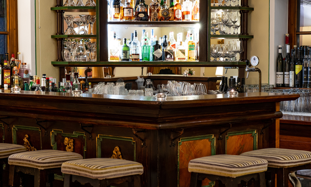 Holzbar mit gestreiften Hockern, Gläsern und Regalen mit verschiedenen Schnapsflaschen und Spirituosen.