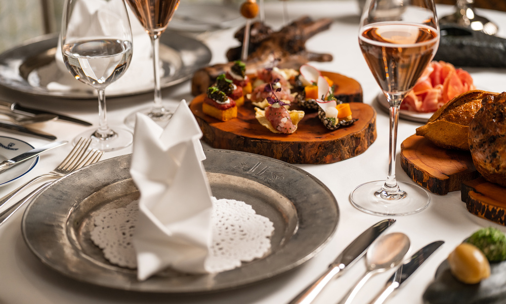 Elegant gedeckter Tisch mit Weingläsern, einer gefalteten Serviette und verschiedenen Häppchen auf Holzbrettern.