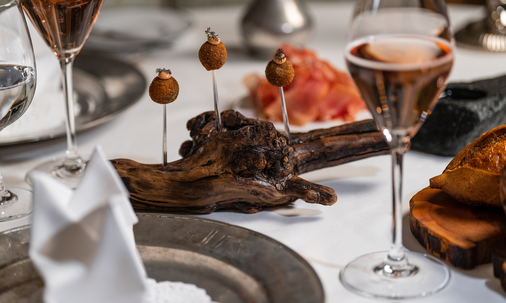 Vier kleine Vorspeisen auf Spießen, die auf einem Stück Holz präsentiert werden, mit Weingläsern und Brot auf einem gedeckten Tisch.