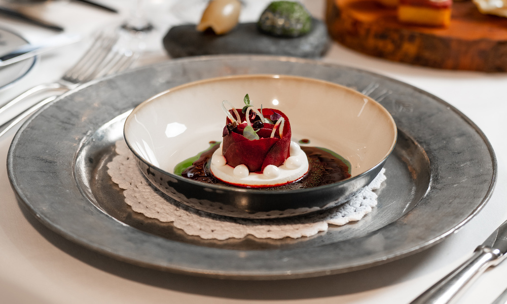 Ein Gourmet-Gericht mit Rote-Bete-Scheiben, Sahne und Garnitur, kunstvoll angerichtet in einer flachen Schale auf einem Spitzendeckchen.