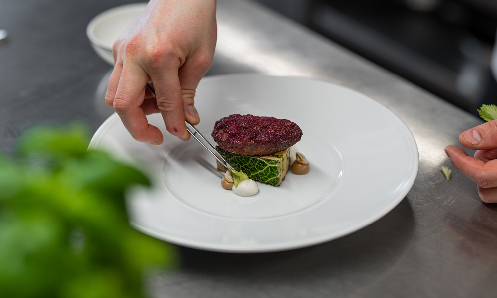 Ein Koch richtet ein Gourmetgericht mit einer Pinzette auf einem weißen Teller in einer Küche an.