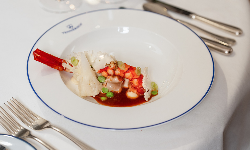 Ein Gourmet-Gericht mit Hummer, Blumenkohl, Erbsen und Soße auf einem weißen Teller, der auf einem formellen Tisch steht.
