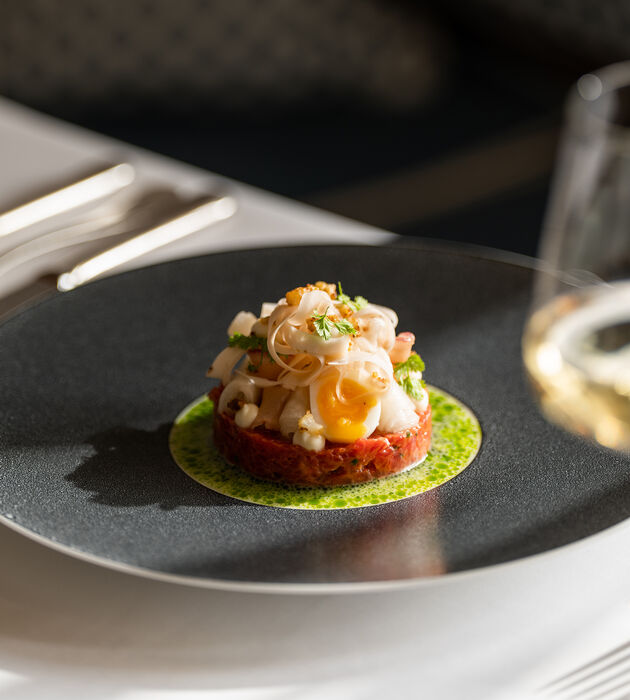 Ein Gourmet-Gericht mit Tartar, Wachtelei und Beilage, kunstvoll auf einem schwarzen Teller angerichtet.
