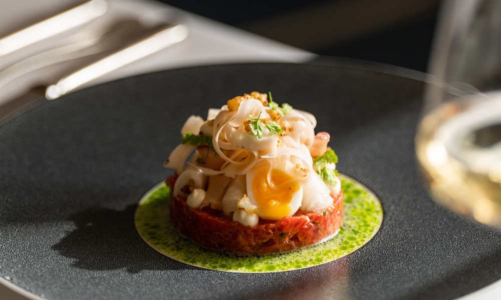 Ein Gourmet-Gericht mit Tartar, Wachtelei und Beilage, kunstvoll auf einem schwarzen Teller angerichtet.