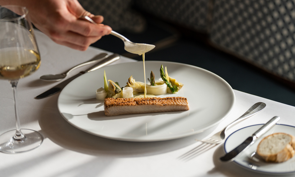 Ein Koch träufelt Sauce über ein Fischgericht mit Spargel und Artischocken auf einem weißen Tischtuch.