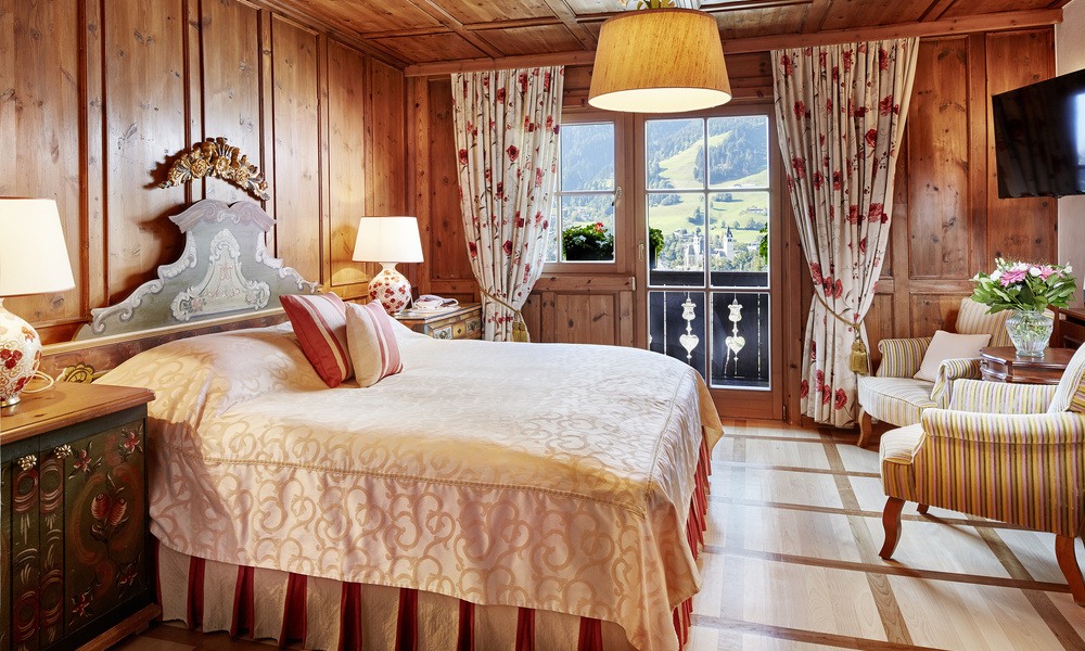 Gemütliches Holzschlafzimmer mit elegantem Bett, geblümten Vorhängen, Sesseln, Lampen und einem Blick auf die Berge draußen.