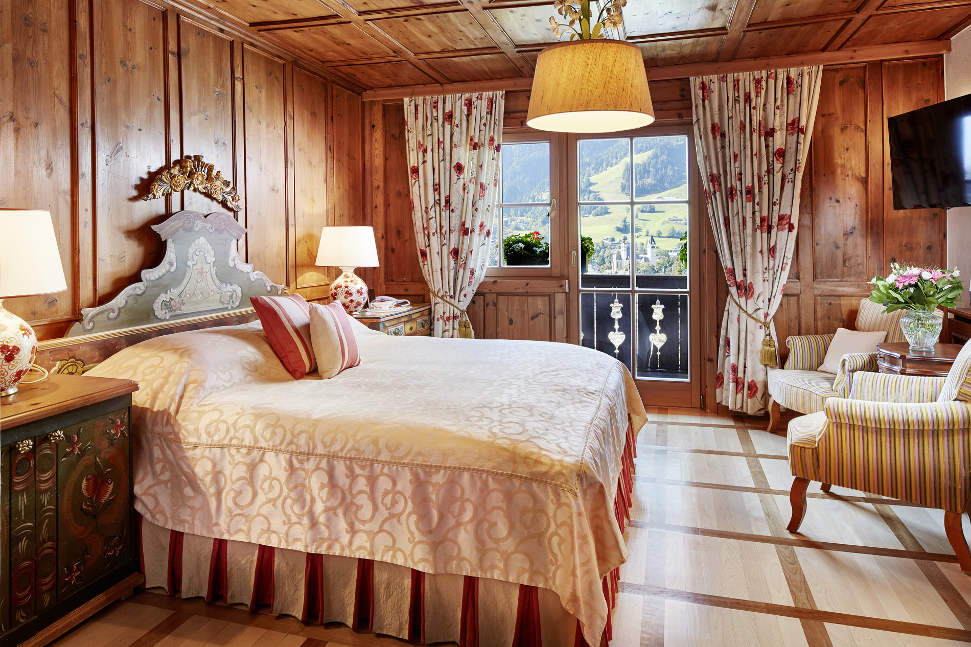 Cozy, rustic bedroom with wood paneling, flowered curtains and a view of the mountains from the window.